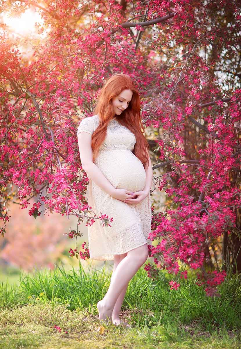 Pregnant woman with red hair posing near cherry blossoms in a beautiful dress.
