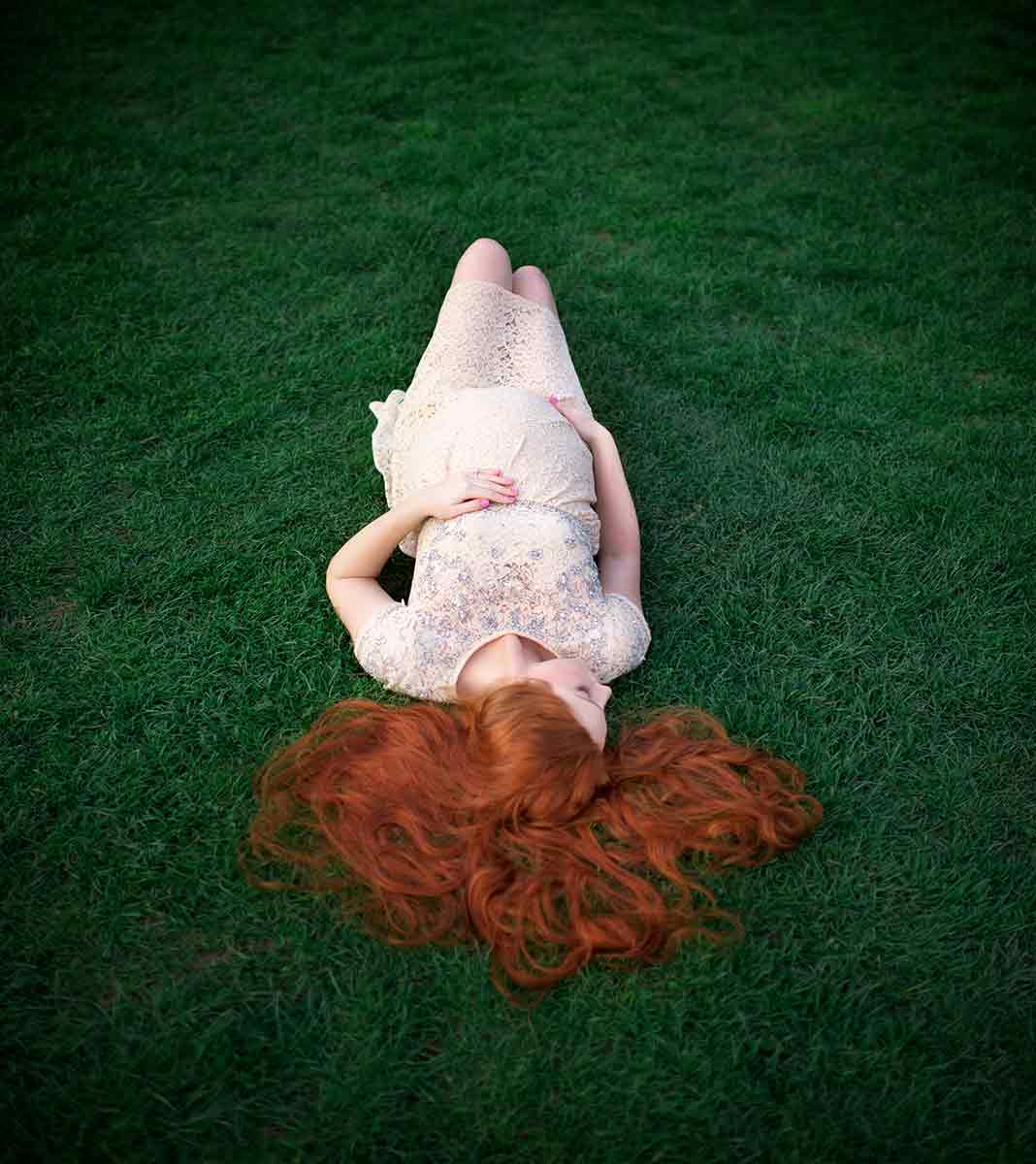 Pregnant woman with red hair laying in grass, cradling her belly.