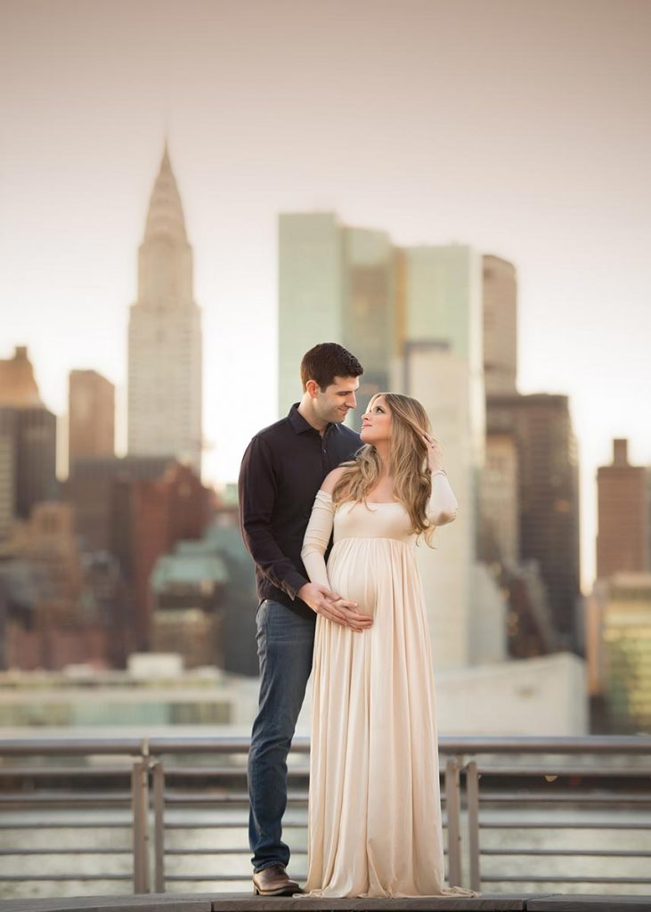 A man holding hands with his wife, and cradling her pregnant belly with the Chrysler building in the back.