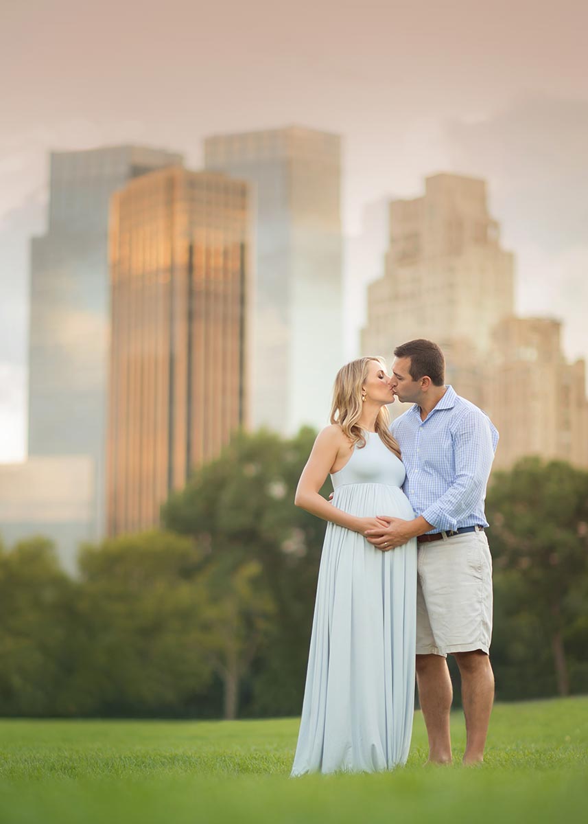 A husband and wife kissing in NYC's Sheep Meadow while cradling a pregnant belly.