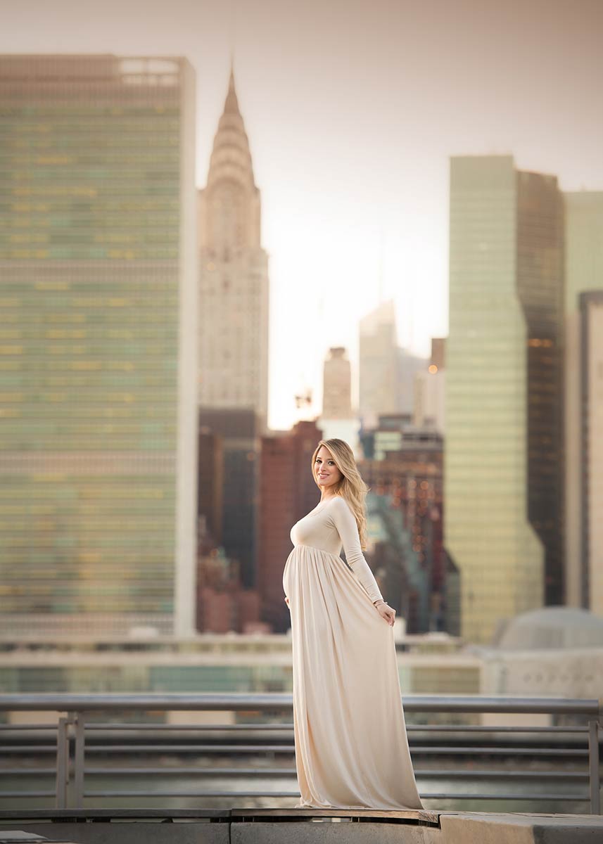 Young blonde woman, posing for a maternity portrait wearing a beige dress with the NYC skyline in the background.