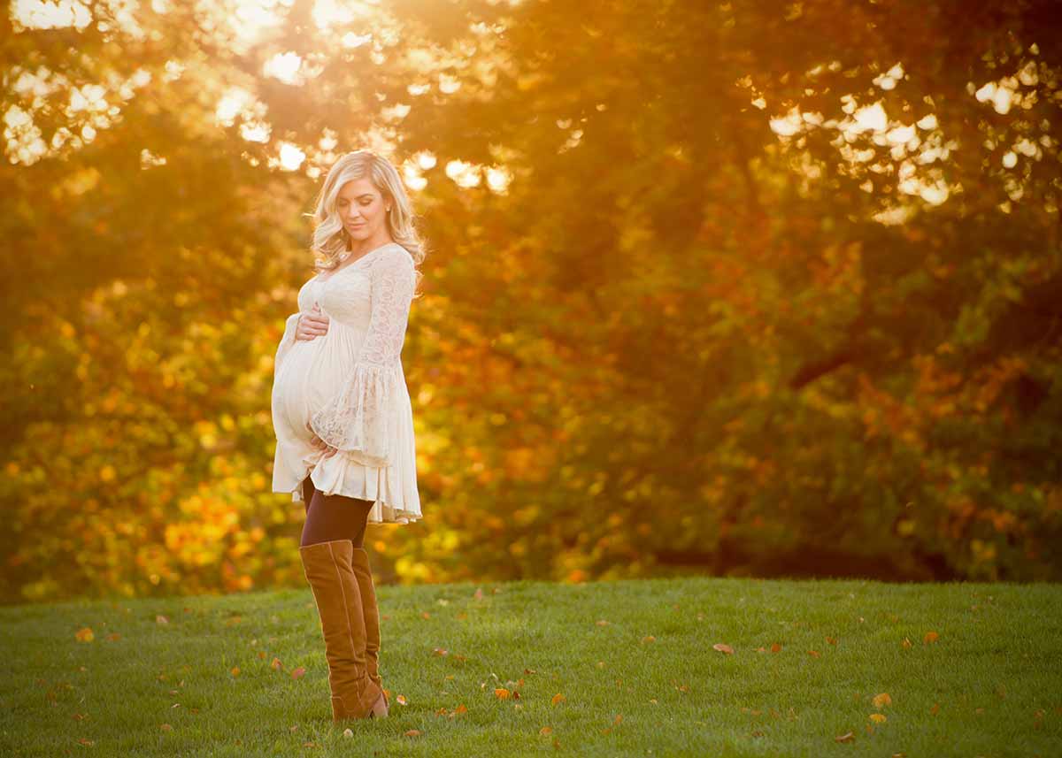 A blonde woman in boots and beige dress cradling her pregnant belly while posing for a maternity portrait in NYC.