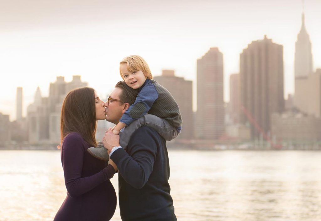 Man and a pregnant woman kissing while holding their sun near NYC's picturesque skyline.