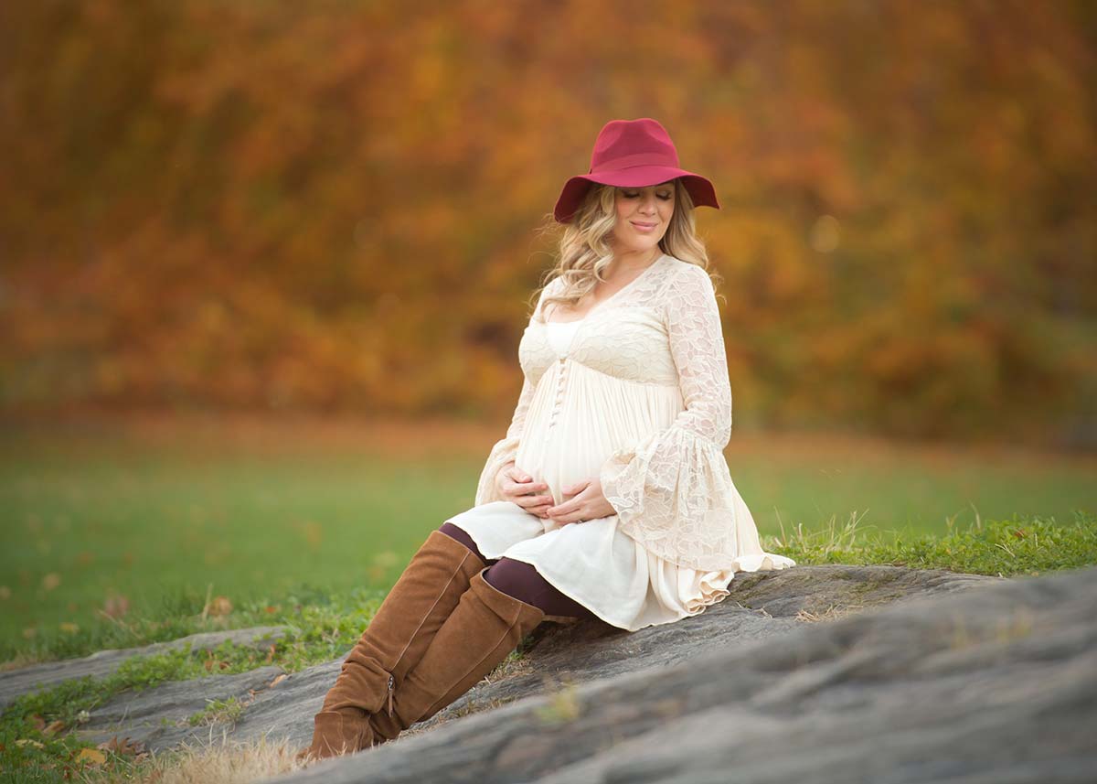 Pregnant blonde woman in a maroon hat and beige dress sitting on a rock in Central Park, NYC.
