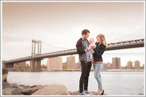 Parents and their baby posing for a family photo with Manhattan Bridge in the background.