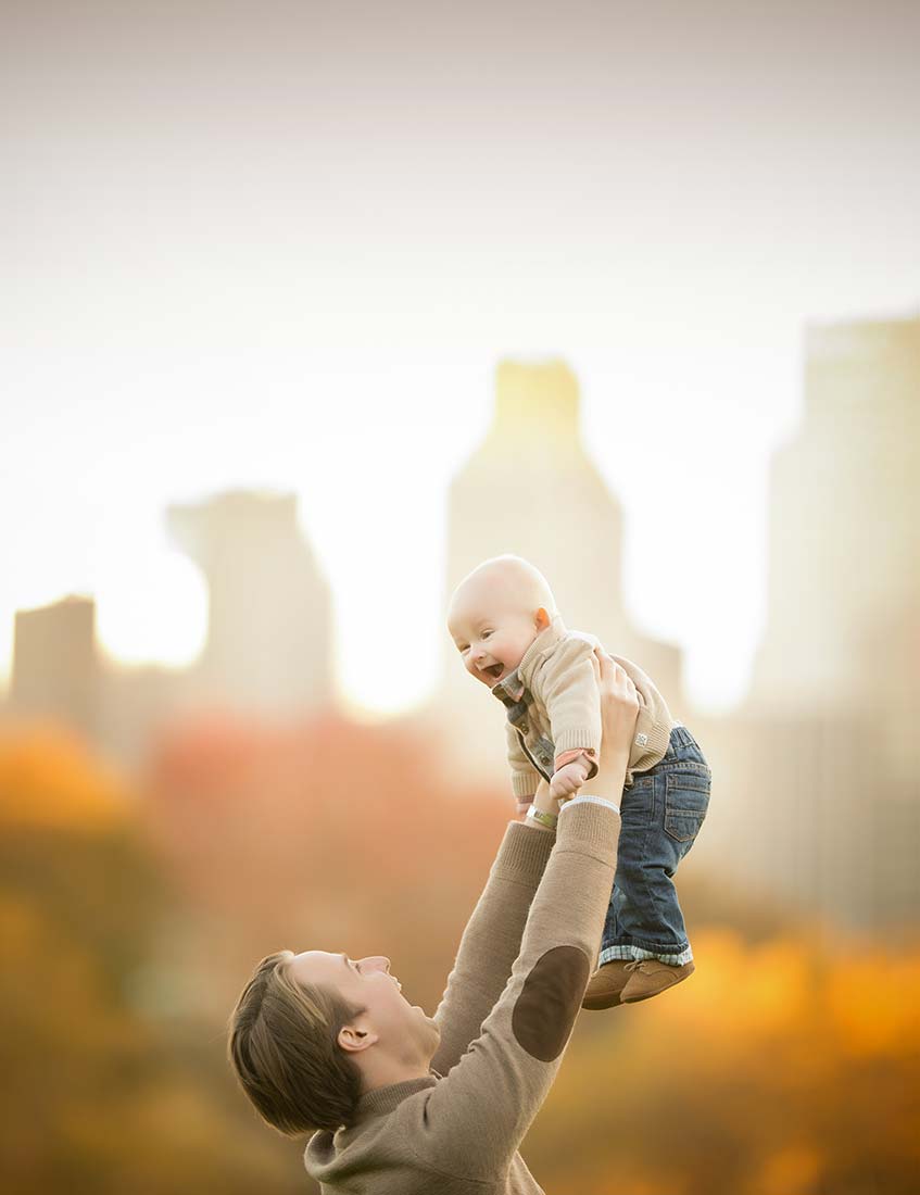 Father holding his baby boy up in the air with NYC skyline behind them.