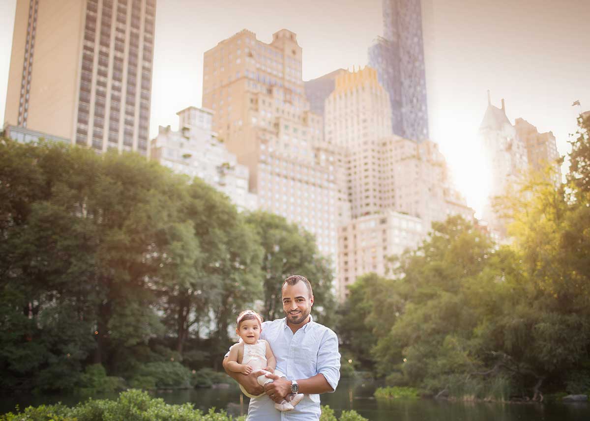 A father holding his daughter pose in front of NYC skyline in Central Park.