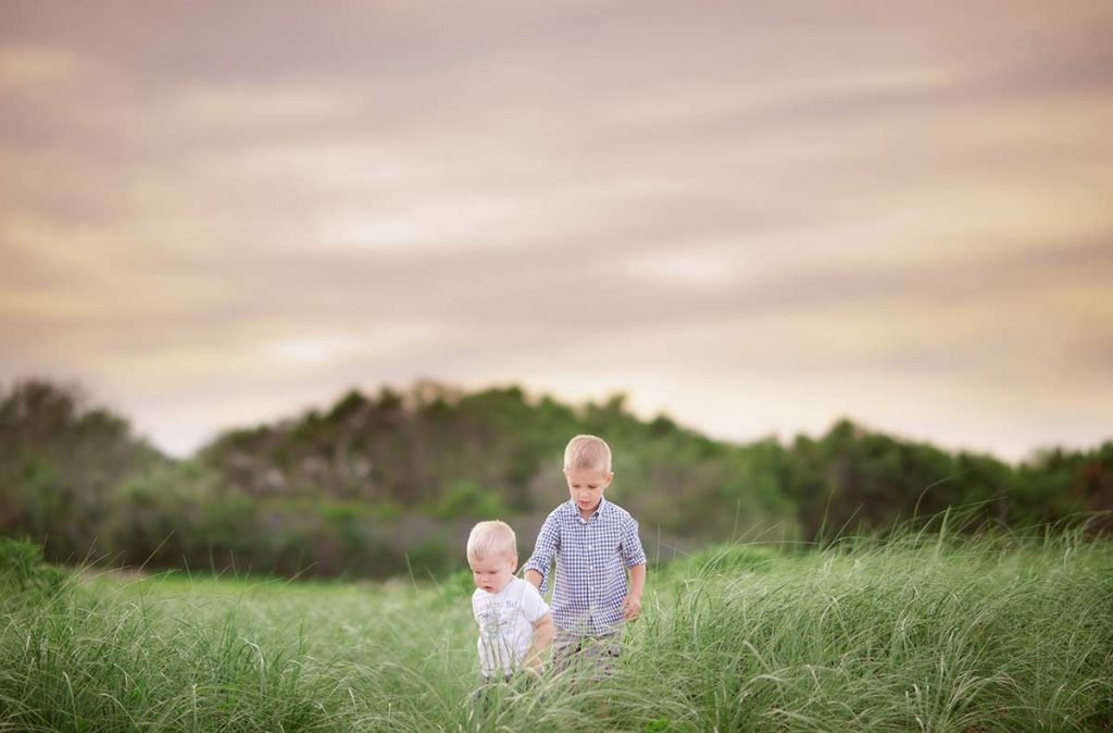 Two young brothers walking through tall grass near a beach.