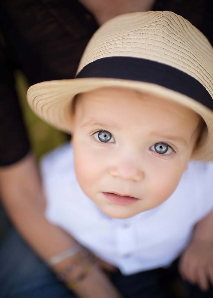 Little boy with blue eyes wearing a fedora hat looking up into the camera.