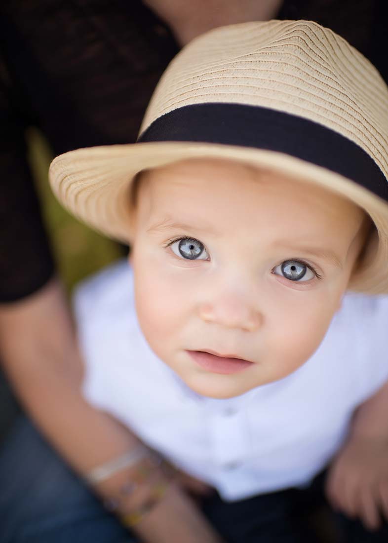 Little boy with blue eyes wearing a fedora hat looking up into the camera.