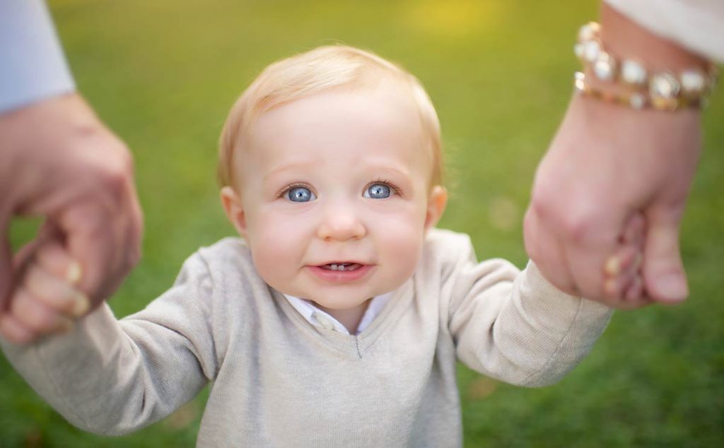 Toddler with blue eyes looking up at the camera while holding his parents' hands.