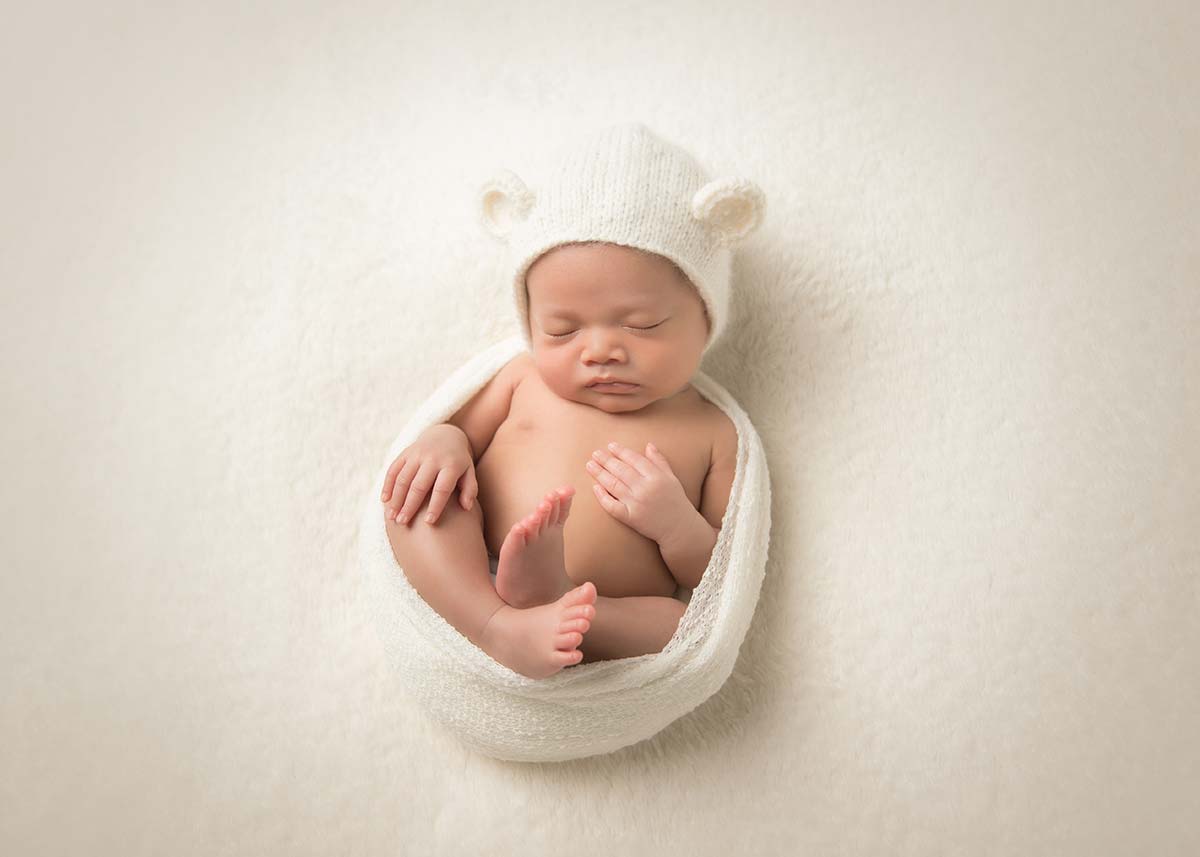 newborn boy sleeping peacefully on his back with a teddy bear hat and cream wrap