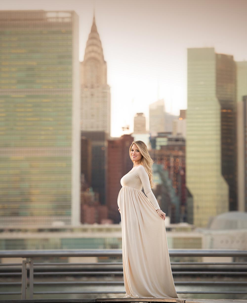 Pregnant blonde woman in a long dress posing in front of the NYC skyline by East River.