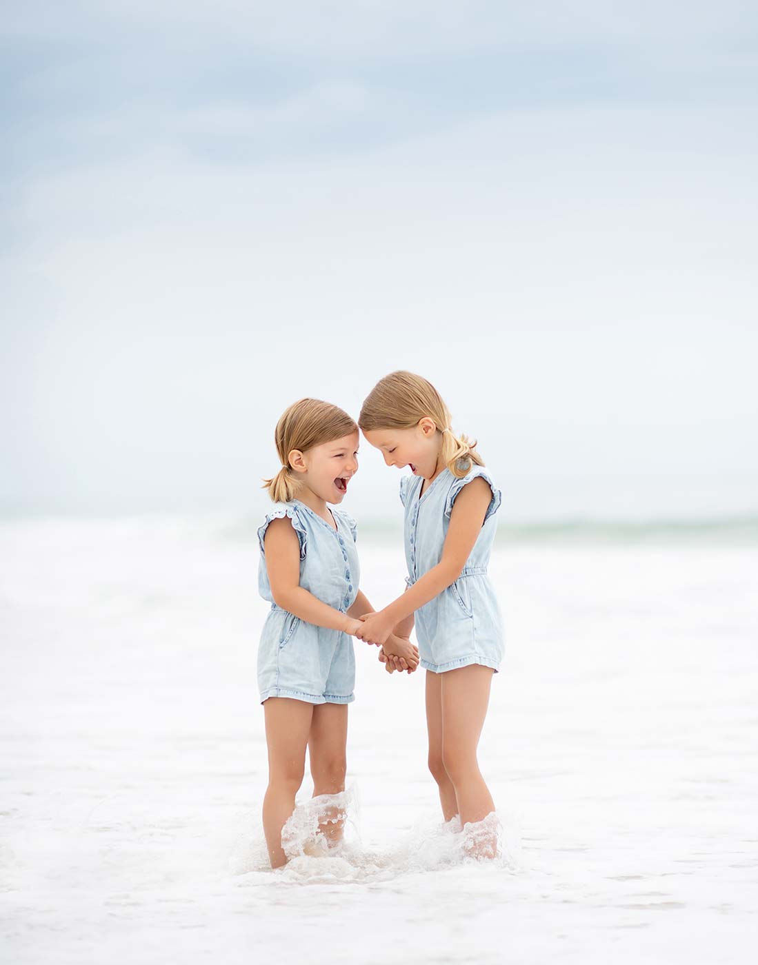 Two sisters in denim dresses sharing a laugh amidst the waves in East Hampton, NY