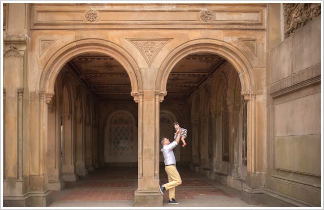 A stylish father holding up his baby daughter in NYC's Bethesda Fountain.