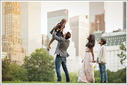 Father holding up his son, while mother and daughter watch in this candid family portrait with NYC skyline as the backdrop.