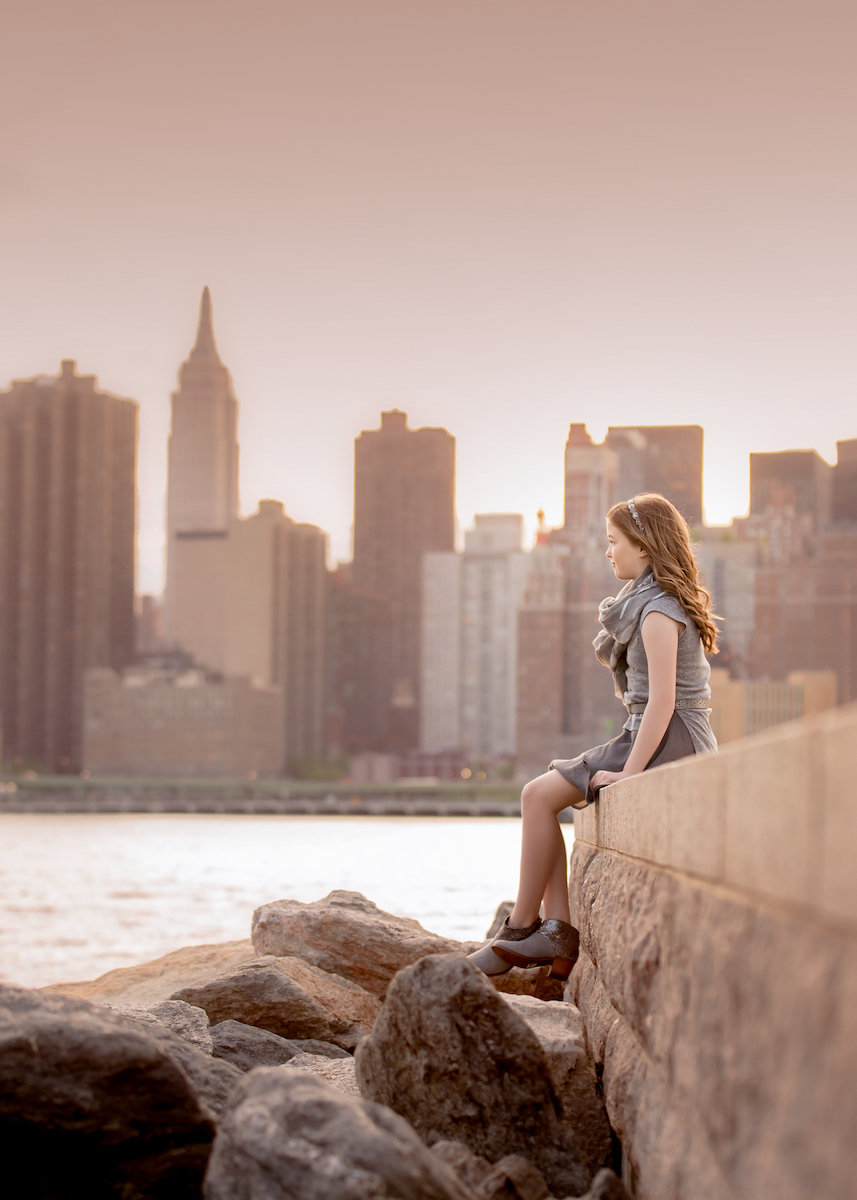 senior portraits sitting on stone wall with nyc skyline backdrop at sunset