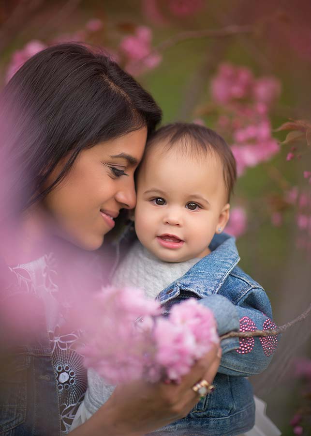 A woman lovingly holds a smiling baby among blooming flowers on the Upper West Side, both in denim jackets and bathed in soft sunlight highlighting their faces.