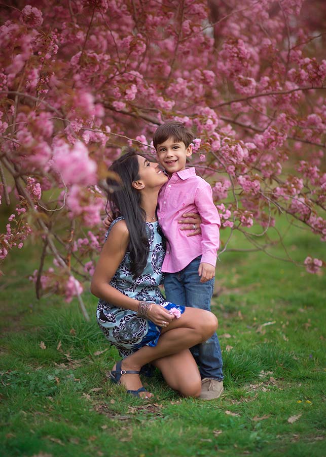 A woman kneels and kisses a smiling young boy on the cheek amid pink flowered trees during a Central Park cherry blossom photoshoot. They are outdoors on green grass, sharing a joyful moment together.