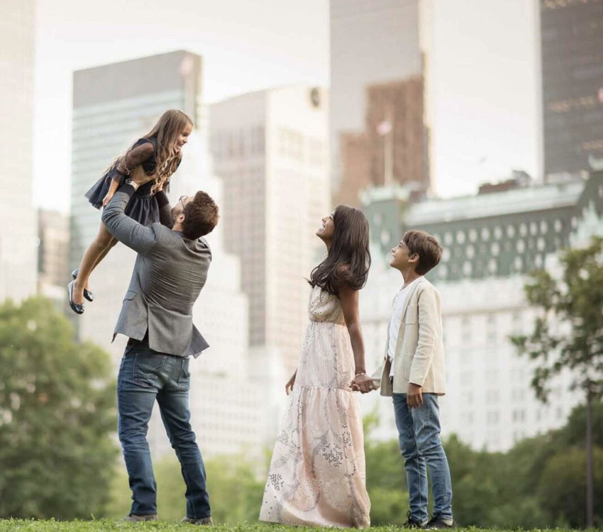 A family of four stands on a grassy area in a city park. A man lifts a young girl into the air. Beside him, a woman in a white dress holds hands with a young boy in a beige jacket. Tall buildings are visible in the background.