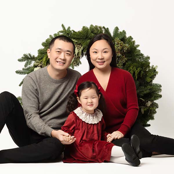 A family of three poses in front of a large green wreath. The man wears a gray sweater, the woman a red sweater, and the child a red dress with a white collar. They are all sitting and smiling at the camera.
