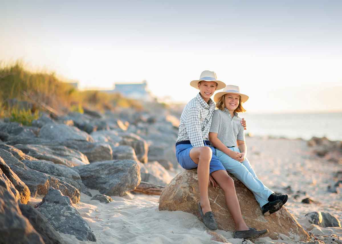 Two brothers in hats and casual clothing sitting on a rock at a sandy beach in the Hamptons, NY during sunset, smiling towards the camera.