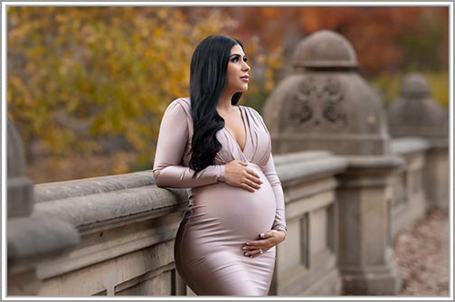 A pregnant woman in a elegant dress leaning on a post in Central Park