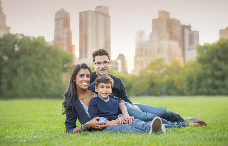 A family of three lies on the grass in a park, with tall NYC buildings visible in the background. A woman with long dark hair, a man with glasses, and a young boy are smiling, facing the camera. The boy wears a navy sweater, and the parents wear casual clothing.