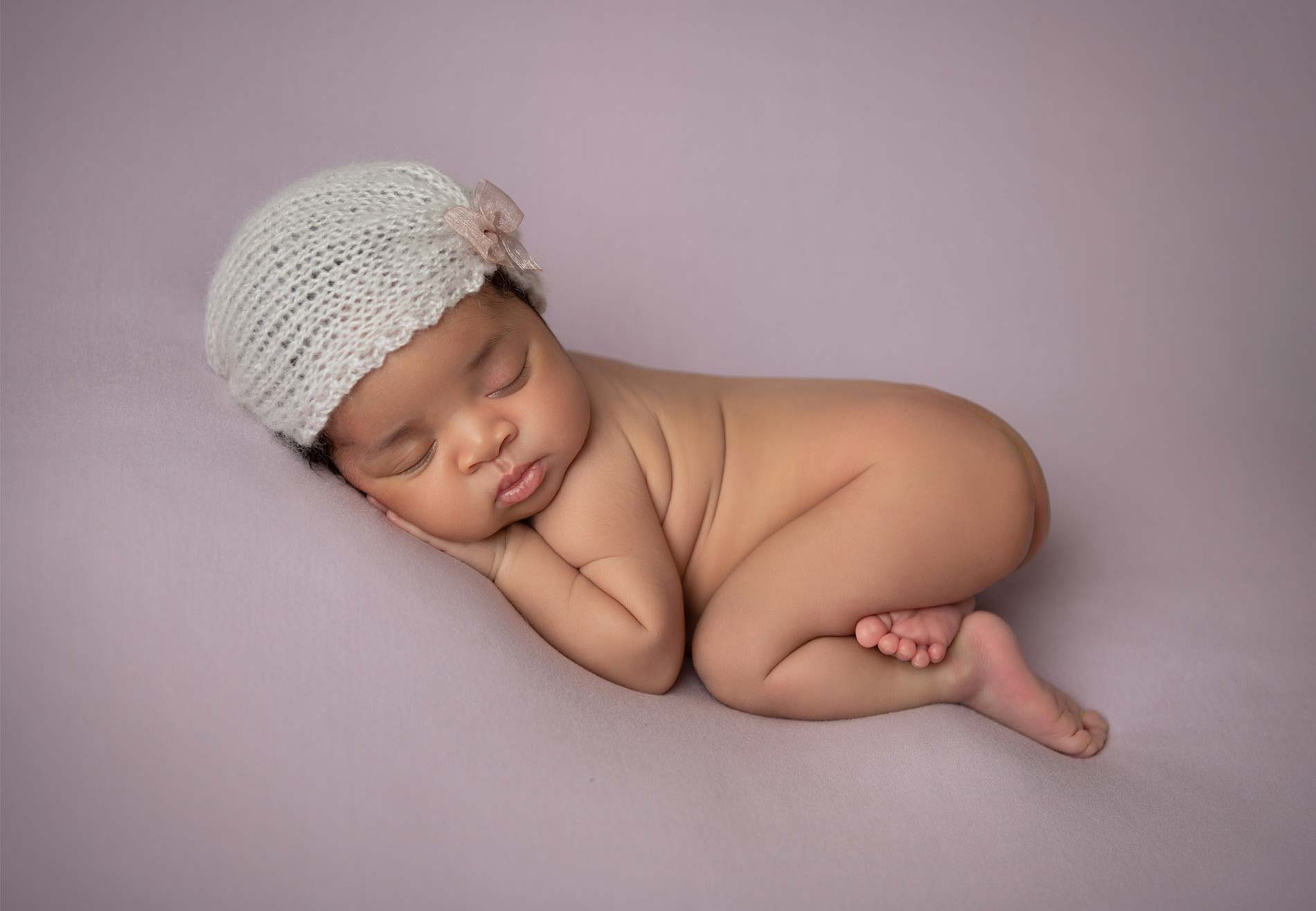 A sleeping baby lies curled up on a soft, light purple surface. The baby is naked, wearing a white knitted cap with a small flower on the side. One arm is tucked under their cheek, and their legs are comfortably bent. Captured by a skilled newborn photographer, the scene is peaceful and serene.