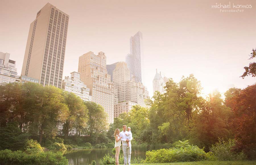 A family of three stands together in Central park with tall buildings in the background. The greenery of the park contrasts with the epic NYC skyline. The photo is taken during sunset, casting a warm, soft light over the scene.