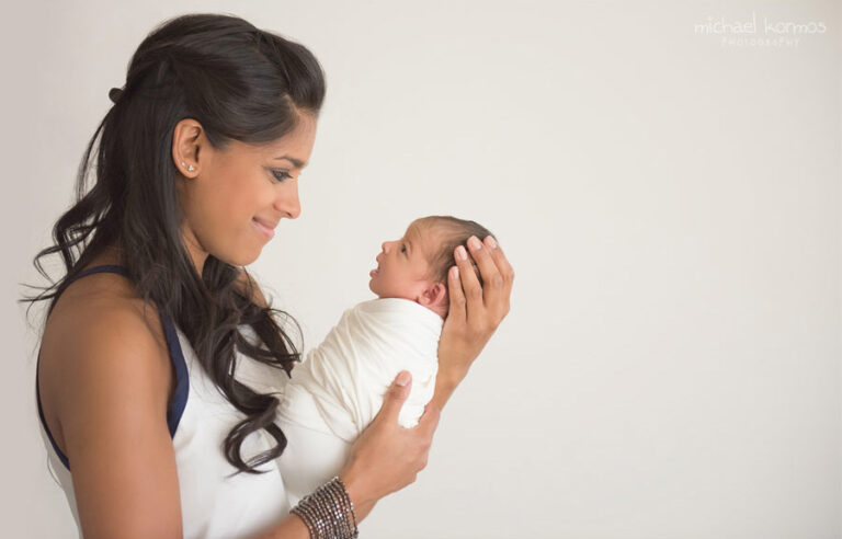 A mother with long dark hair, wearing a sleeveless top, lovingly cradles her newborn baby swaddled in a white blanket. They are facing each other, and the mother is smiling gently at the baby. The background is plain and light-colored.