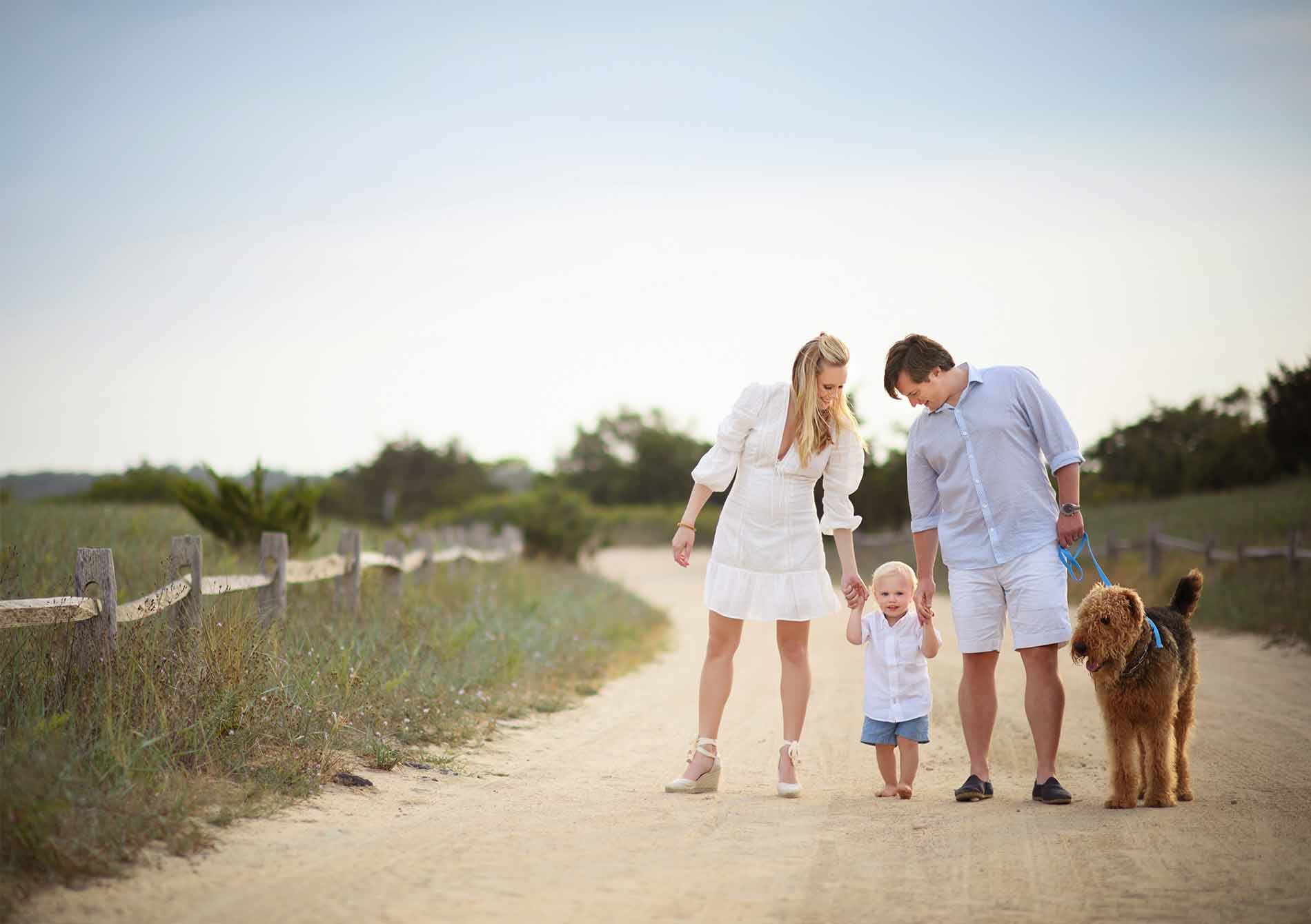 A Hamptons photographer captures a family walking down a dirt path lined with wooden fences and greenery. The parents, dressed in light-colored outfits, hold hands with their young child in the middle. They have an orange-brown dog on a blue leash walking beside them under the clear, blue sky.