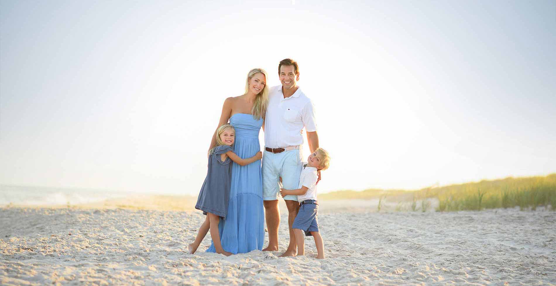 A family of four smiles and stands on a sandy beach. The mother, wearing a blue dress, and the father, in a white shirt and light blue shorts, embrace their young daughter and son, who both wear blue outfits. The sky is clear and the grass dunes are visible in the background.