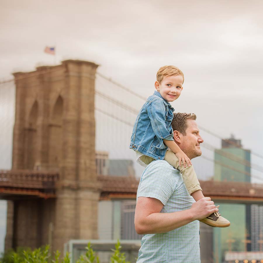 Boy sitting on his father's shoulders with NYC's iconic Brooklyn Bridge in the background