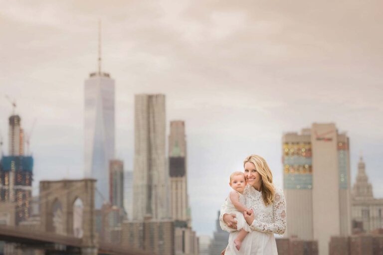 A photographer captures a smiling mother in a white lace dress holding her daughter, with skyscrapers including One World Trade Center and the Brooklyn Bridge as their backdrop. The sky is overcast.
