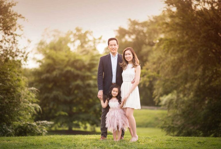 A family of three, consisting of a man, a woman, and a young girl, stands on a grassy area against a backdrop of lush greenery. The man and woman are dressed in formal attire, while the girl wears a light pink dress. Captured by an expert family photographer from NYC, the scene is lit by soft, natural light.