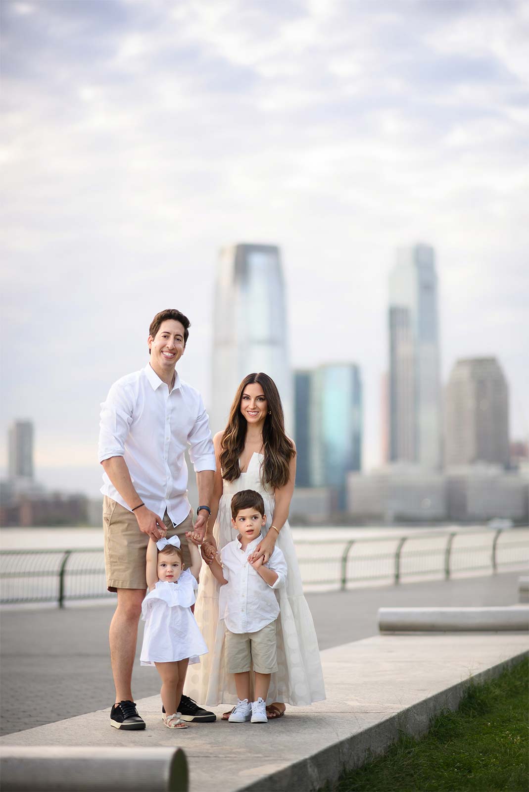 A family of four poses outdoors in front of the NYC waterfront skyline. The father and mother stand on either side, each holding the hand of a young child. The father and two children wear white shirts and beige shorts, while the mother wears a white dress.
