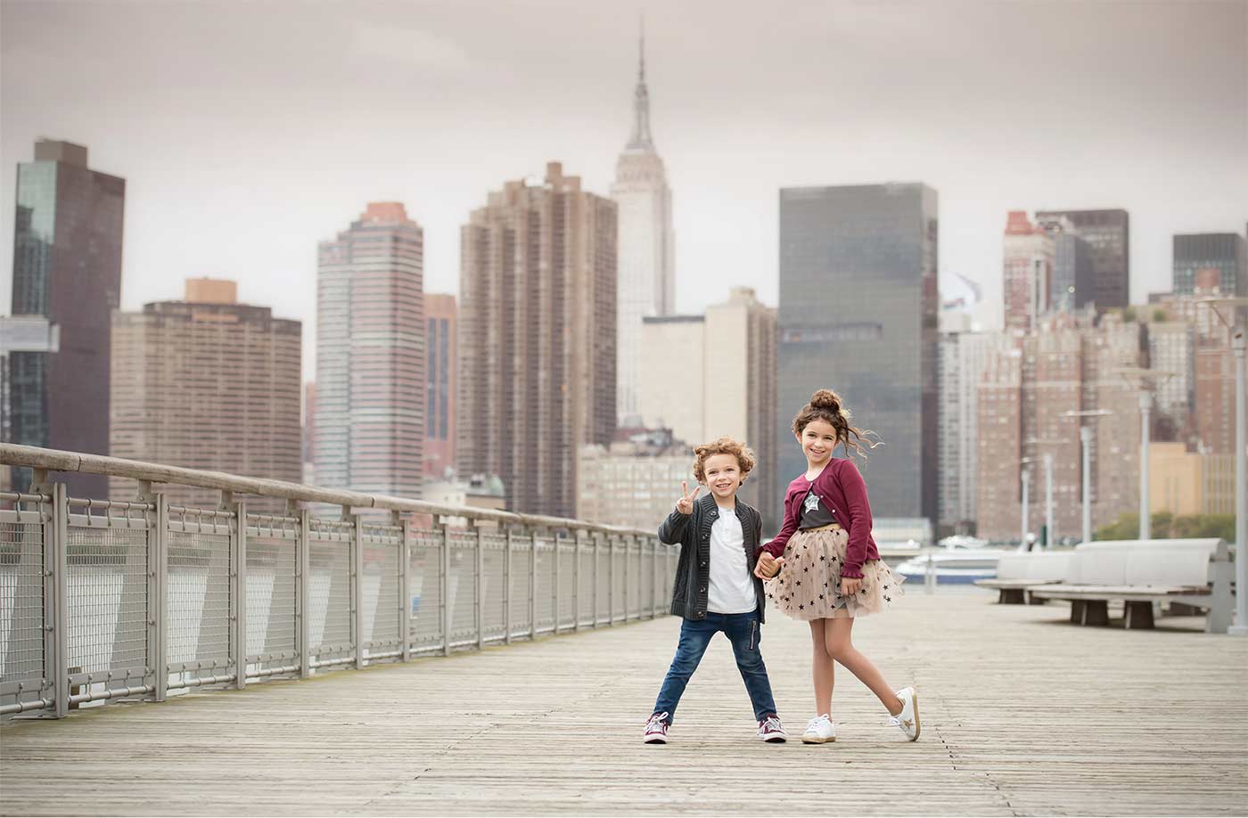 Two siblings stand on a wooden pier with the NYC skyline in the background. One holds up a peace sign while the other has one hand on their hip. Tall buildings, including the Empire State Building, rise against a cloudy sky.