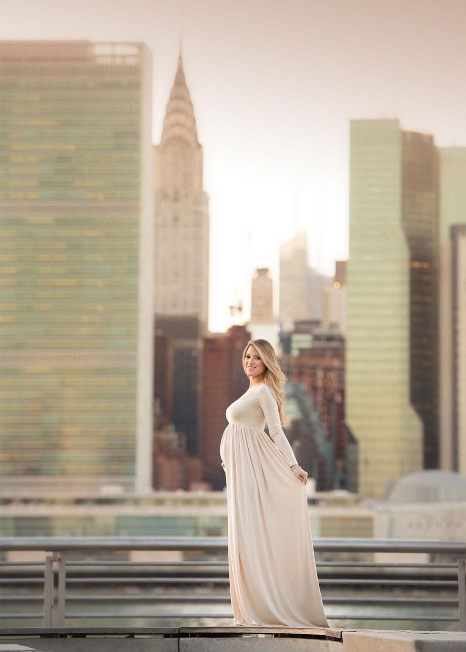 A pregnant woman in a flowing cream-colored dress stands on a terrace with the NYC skyline in the background, including notable skyscrapers. She is smiling and holding her dress, highlighting her baby bump. The scene is bathed in soft, warm light.