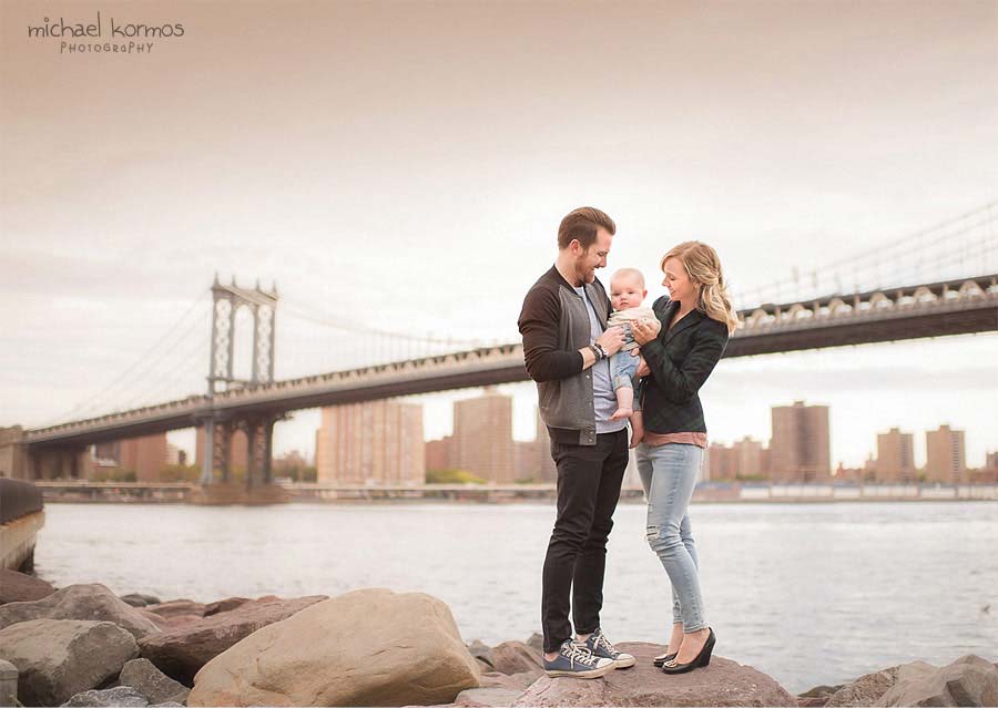 A man and a woman stand on rocks near the water, holding a baby. The Manhattan Bridge, along with the city skyline, are visible in the background. The man wears a black jacket and jeans, while the woman wears a black jacket and blue jeans. The baby is bundled in a white outfit.