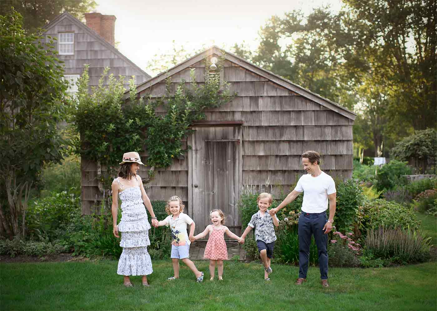 A family of five, including two adults and three young children, is holding hands and walking in front of a rustic wooden shed surrounded by greenery. The adults wear summer clothes, with the woman in a white dress and hat, and the man in a white shirt and jeans—capturing the essence of a Hamptons family getaway.