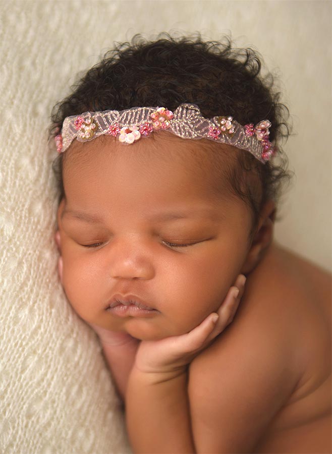 A close-up of a sleeping baby with curly hair resting on a soft surface. The baby is wearing a delicate headband adorned with flowers and lace. Their hands are gently tucked under their chin. The background is textured and light-colored.