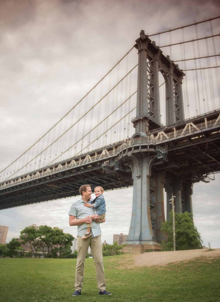 A man holding a child stands on a grassy area beneath the Manhattan Bridge. The cloudy sky and steel structure of the bridge create a dramatic backdrop. Trees and buildings are visible in the background.
