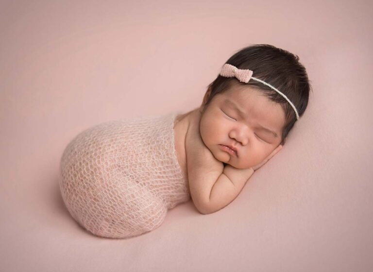 A newborn baby is peacefully sleeping on a soft pink blanket. The baby is wrapped in a delicate pink knit fabric and is wearing a small pink bow headband. The baby's head is resting on its arm, and the overall scene exudes calmness and innocence.