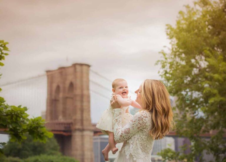 A mother with long blonde hair, wearing a white lace dress, holds her smiling daughter in a light-colored dress. They are outdoors with the Brooklyn Bridge and a cloudy sky in the background. Surrounding greenery frames the scene.