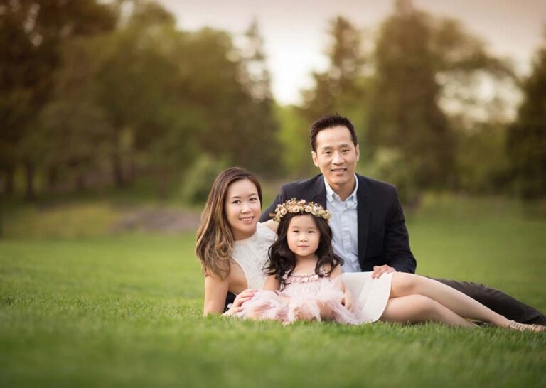 A family sits on the grass in an outdoor setting of Central Park. A woman with long hair is on the left, a man in a dark suit is on the right, and between them is a young girl wearing a dress with a floral headband and tulle skirt. Trees and greenery are blurred in the background.
