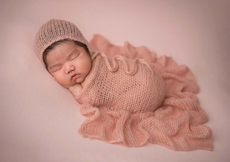 A peacefully sleeping baby is wrapped in a soft, knitted pink blanket with a matching knit hat. Captured by a talented newborn photographer, the infant lies on a light pink surface, enhancing the gentle and serene atmosphere of the image.