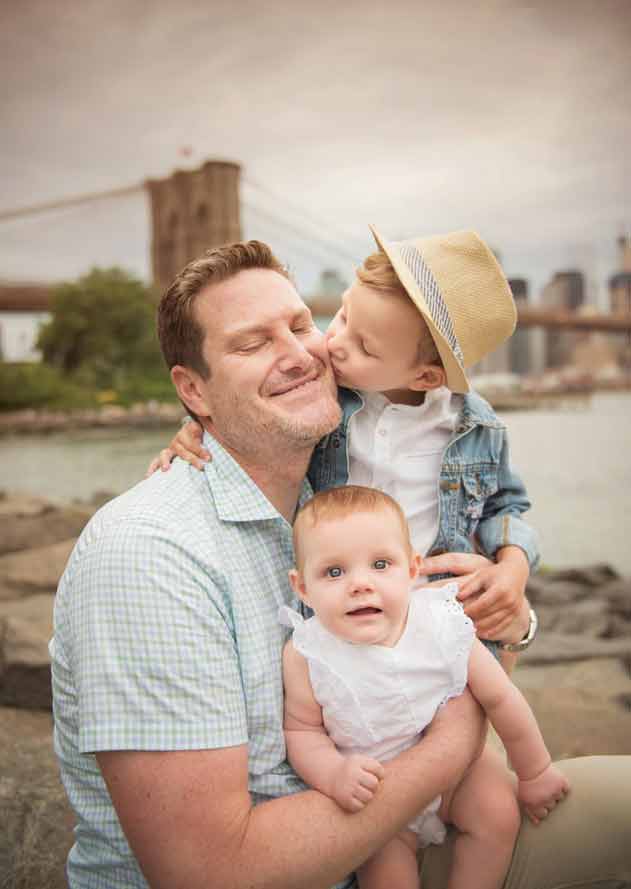 A father, smiling warmly, holds a baby in his arms while a young child, wearing a hat and denim jacket, kisses him on the cheek. They are outdoors near water with a bridge and city skyline in the background.