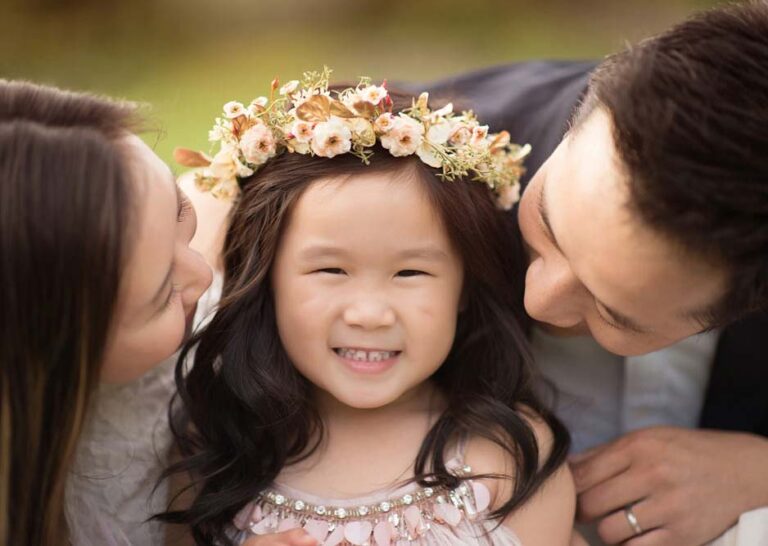 A young girl wearing a floral crown smiles joyfully at the camera, flanked by two adults, one on each side, who are looking at her lovingly. The background is softly blurred, highlighting the affectionate and happy moment shared among them.