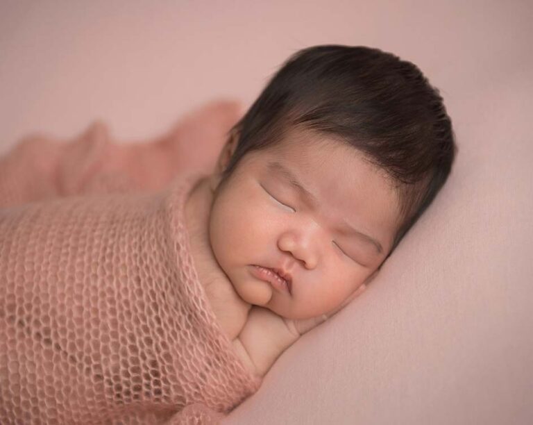 In an NYC photo studio, a newborn baby with closed eyes is sleeping peacefully on a soft pink blanket. The baby is wrapped in a delicate pink knit shawl, with their tiny hands tucked under their cheek. The background is also pink, creating a warm and gentle atmosphere.